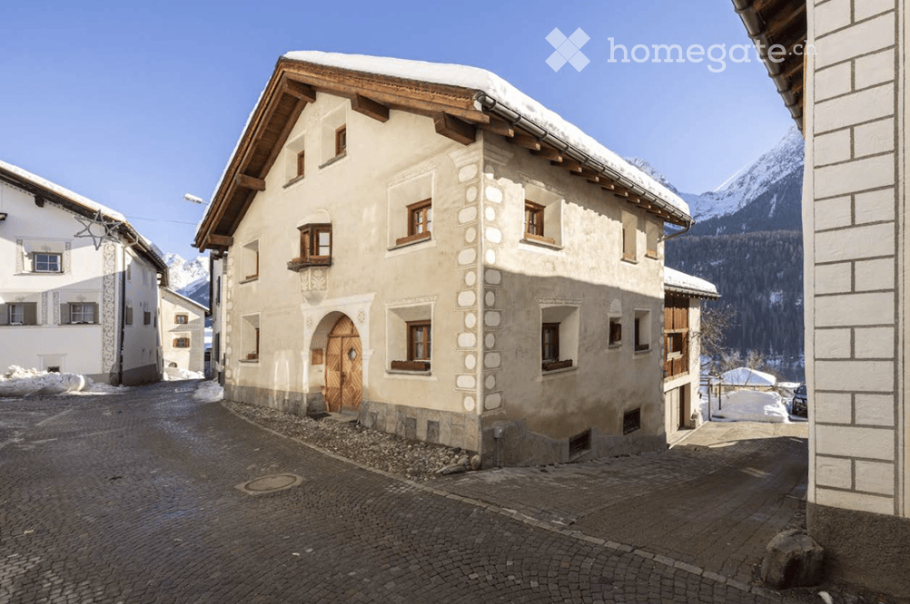 Traditional alpine building with a wooden roof, stone facade, and arched doorway, situated in a snow-covered village with mountain views.