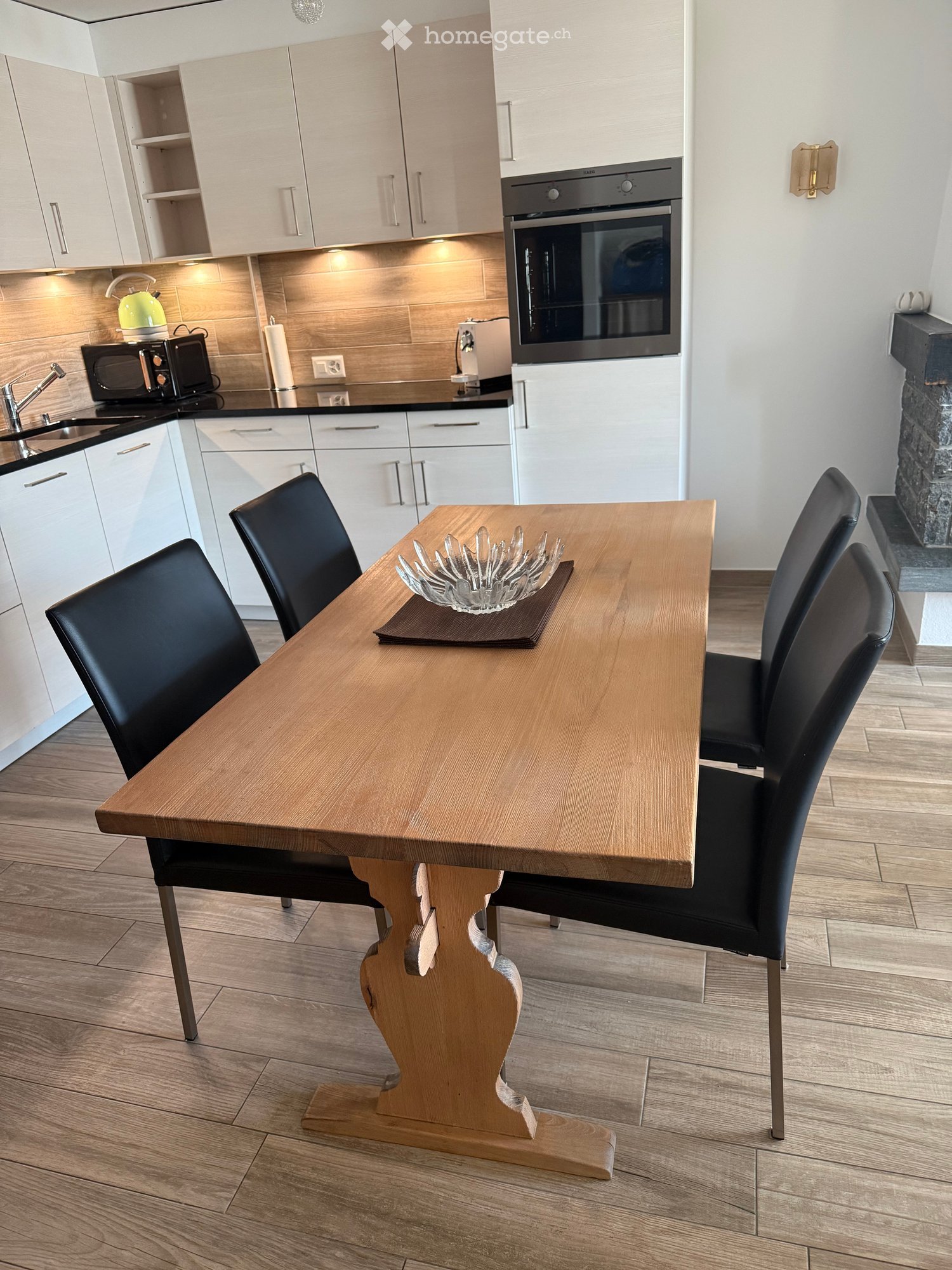Modern kitchen with white cabinets, a wooden dining table with four black chairs, an oven, microwave, and a teapot on the counter.