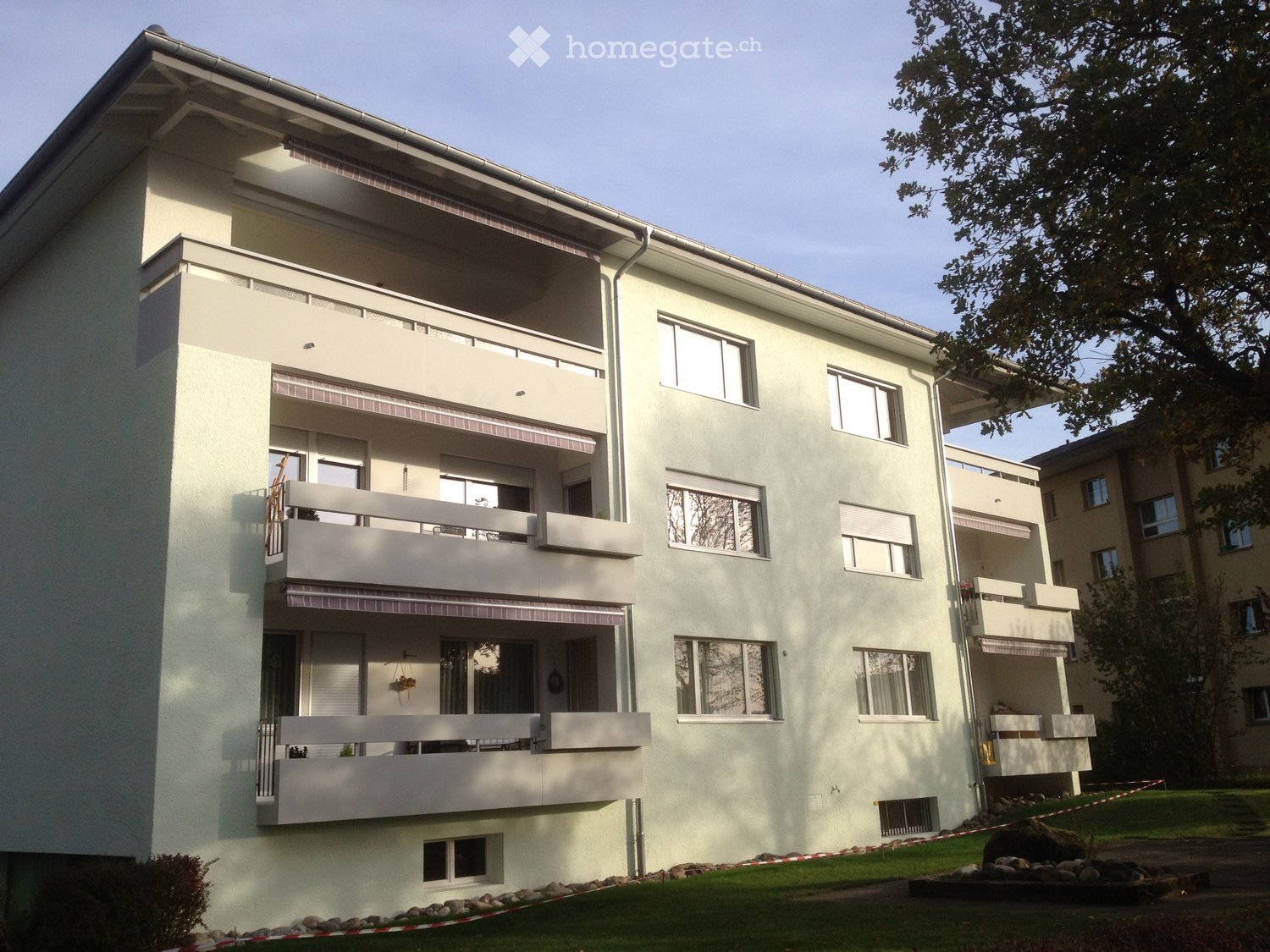 Modern three-story residential building with white exterior, multiple balconies, and large windows. The building features a clean, contemporary design with a flat roof and is surrounded by a well-maintained lawn and small garden area.