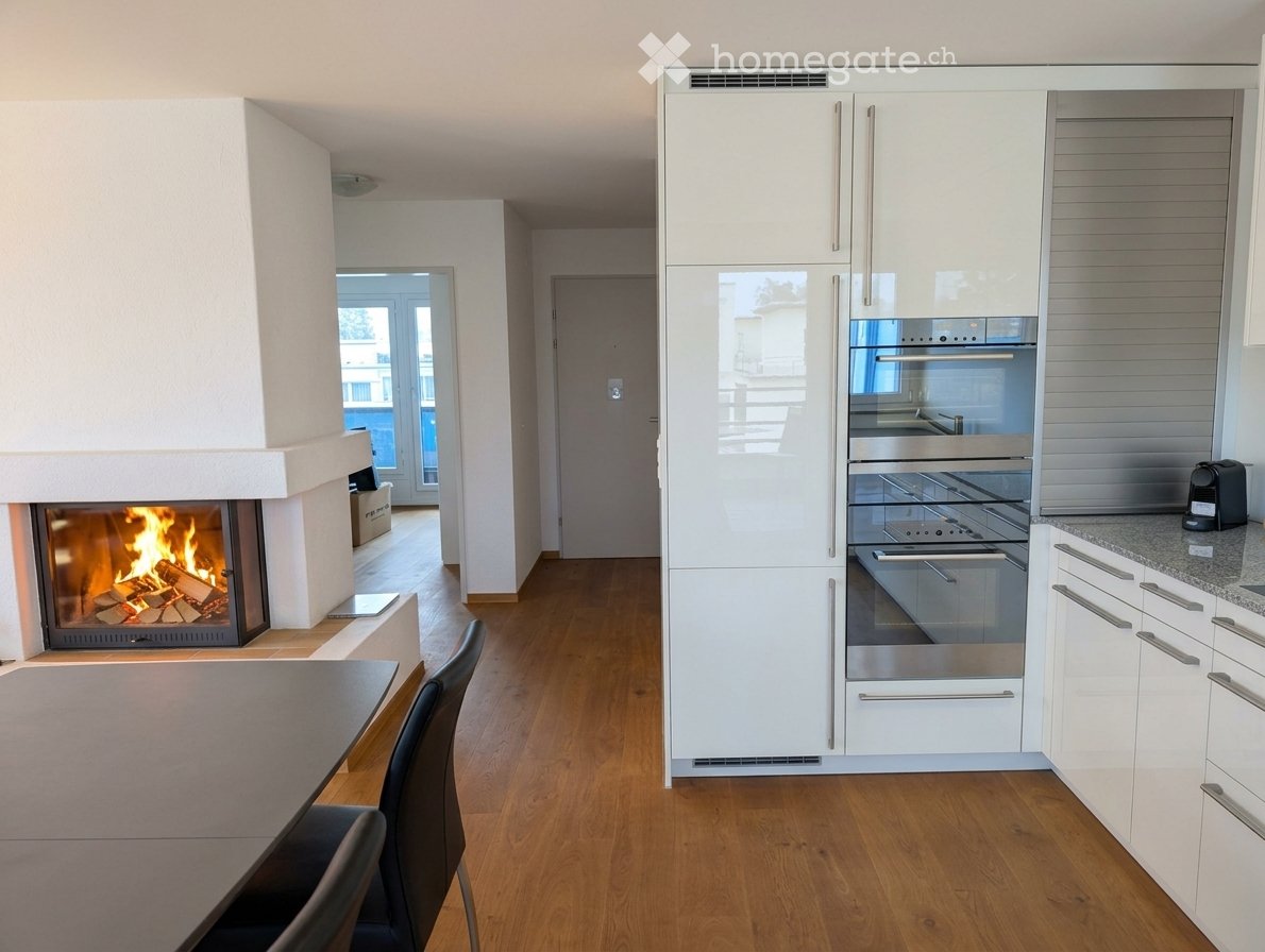Modern kitchen with white cabinets, stainless steel appliances, a built-in oven, and a fireplace in the adjacent living area. The space features a wooden floor and a dining table with chairs.