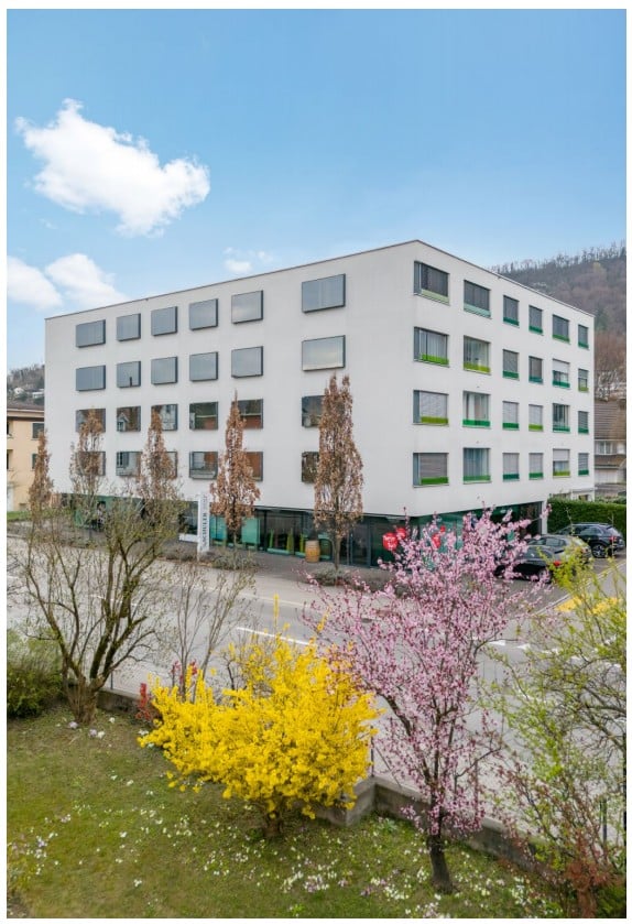 Modern multi-story building with white facade and green window frames, surrounded by spring flowers and trees, with parked cars visible in front.