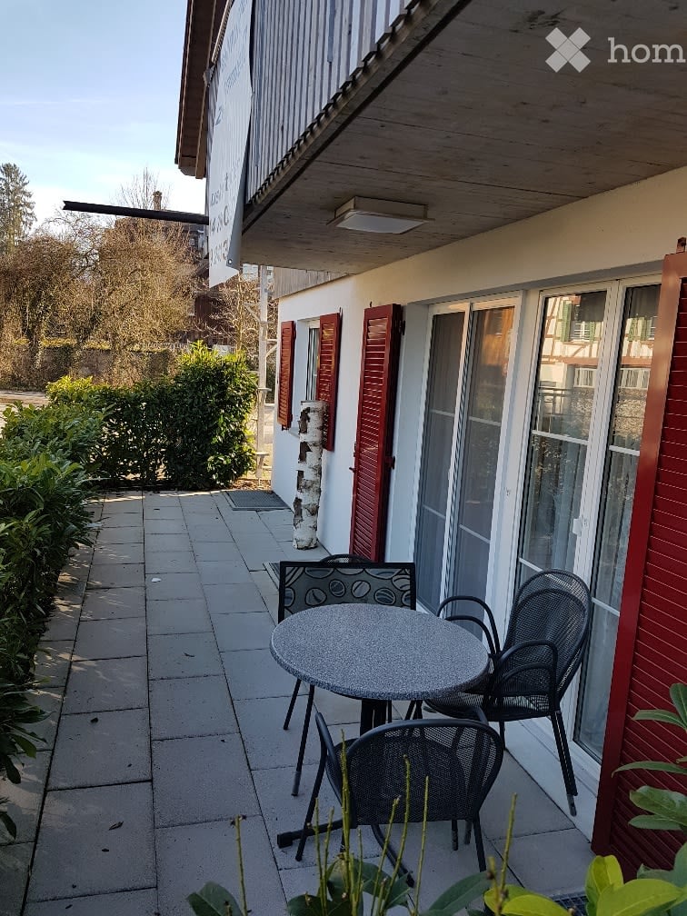 Balcony with a round table and two black metal chairs, surrounded by greenery and a building with red shutters.