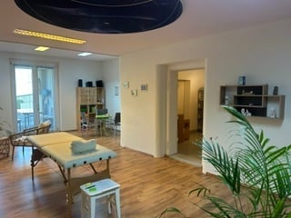 Bright, modern room with wooden flooring, massage table, chairs, small stool, shelves, and a potted plant near the window.