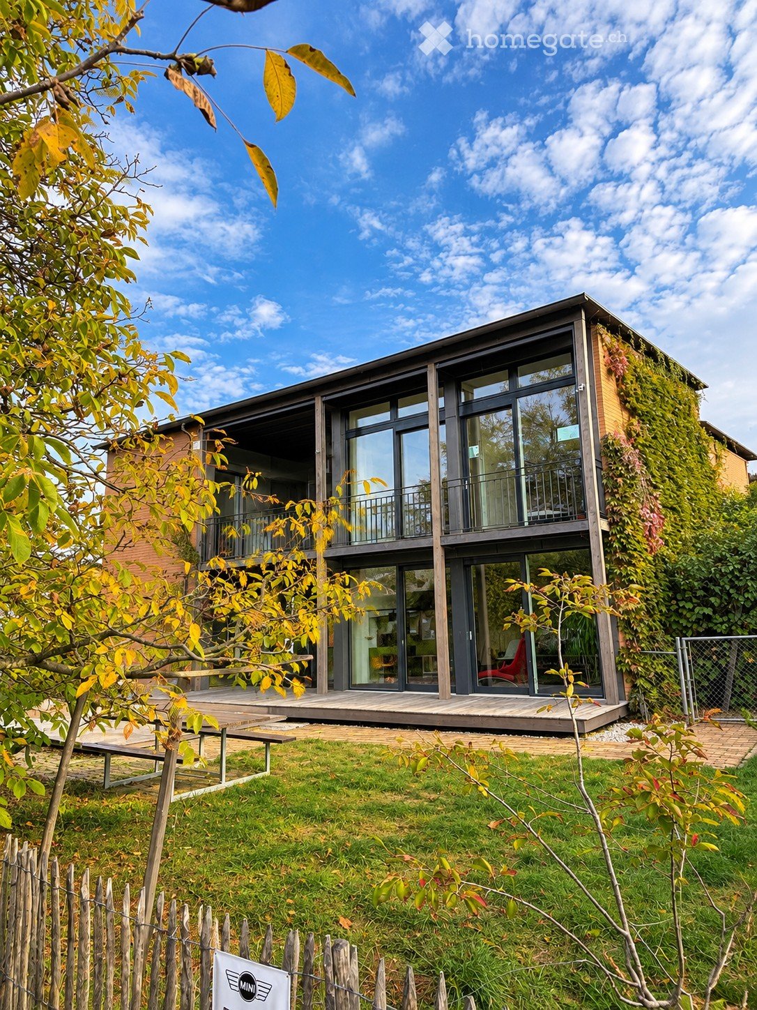 Modern two-story house with large glass windows, a balcony, and a green living wall on one side. The house is surrounded by a well-maintained garden with a wooden fence and a small patio area.