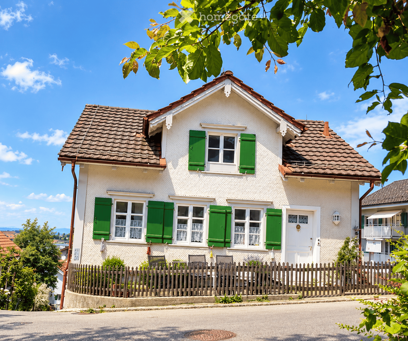 A two-story white house with green shutters and a red-tiled roof, surrounded by a wooden fence and garden area.