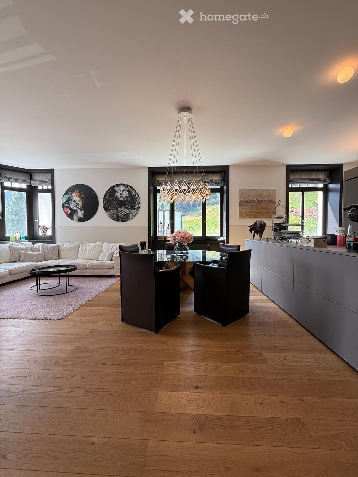 Open-plan living and dining area with light wooden flooring, white sofa, glass dining table, modern chandelier, and large windows.