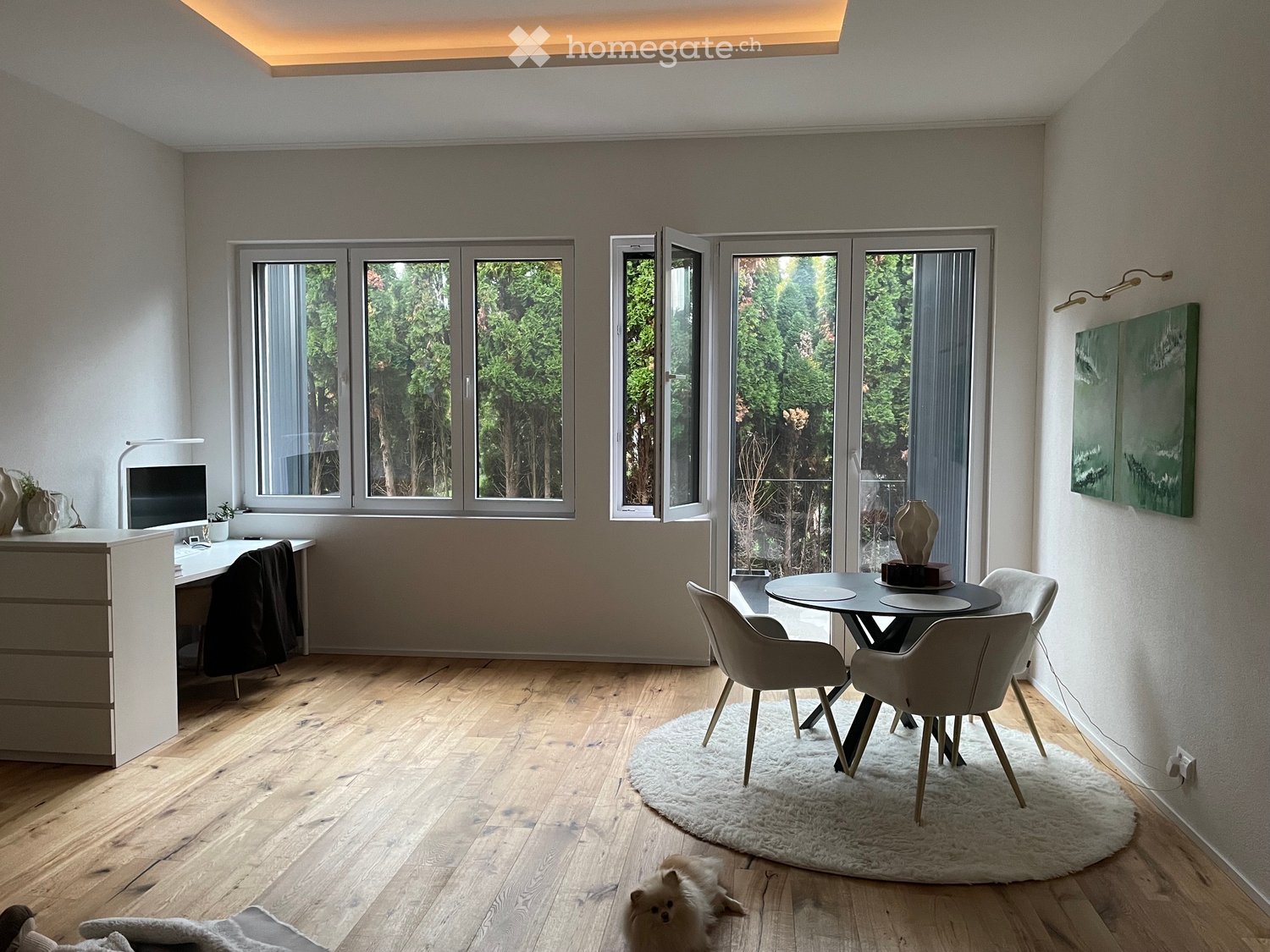 Modern, well-lit room with wooden flooring, large windows, a desk with a screen, a round dining table with chairs, and decorative vases.