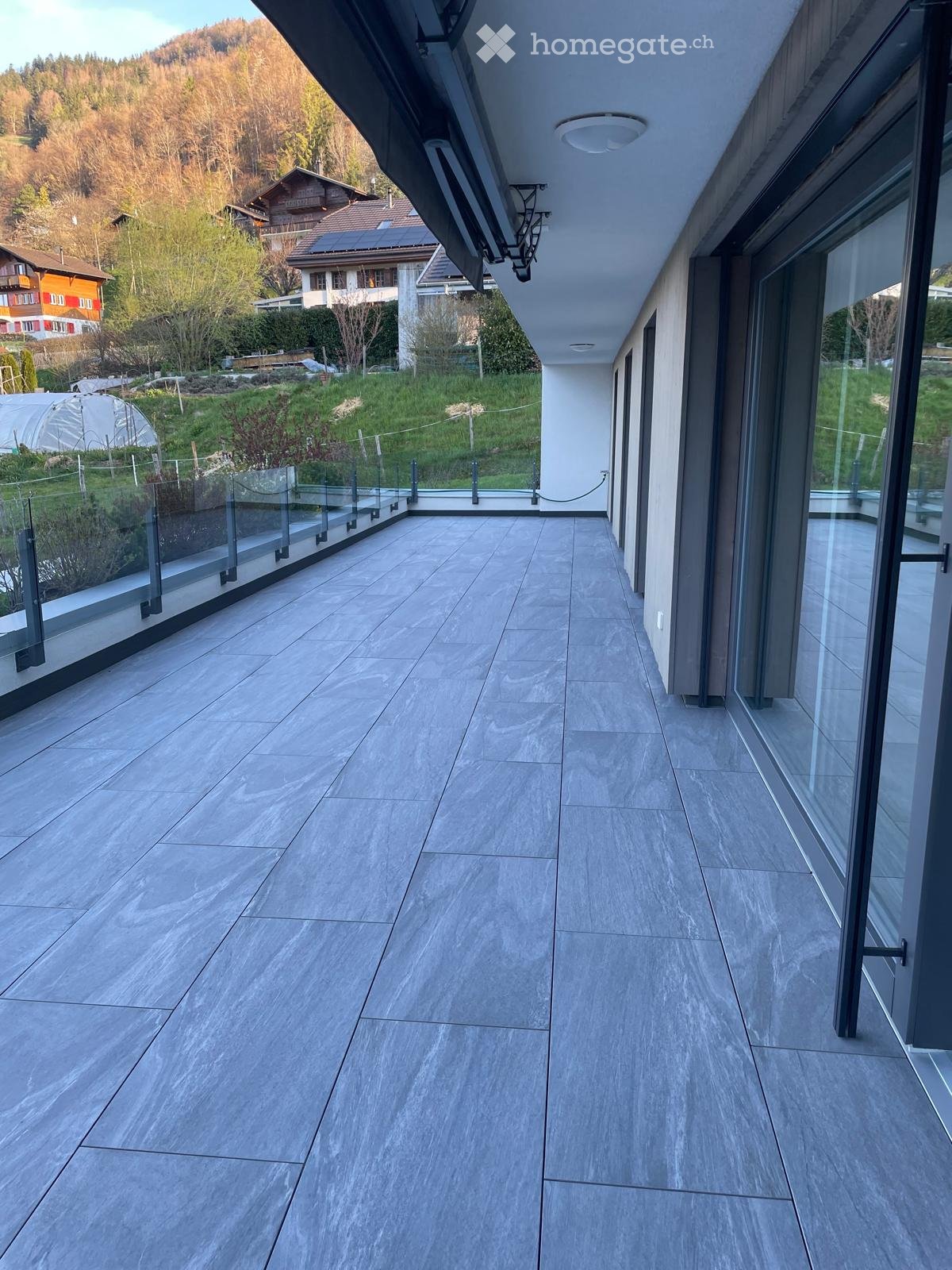 Modern balcony with grey tiled flooring, glass railings, and a view of a landscaped garden and surrounding houses.