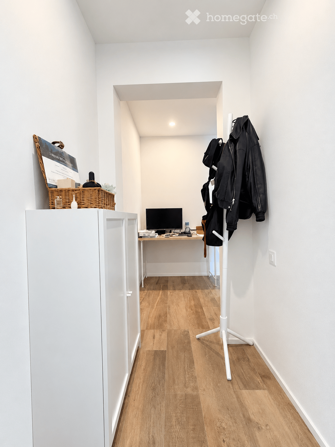 A narrow hallway with wooden flooring, a small desk with a computer, a coat rack with jackets, and a wicker basket on top of a cabinet.