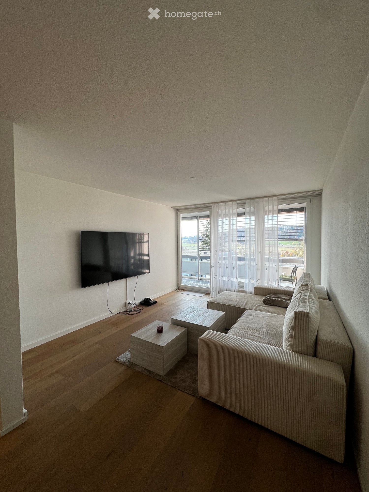 Living room: beige sectional sofa, glass coffee tables, wall-mounted TV, windows with white curtains, white walls, hardwood floors