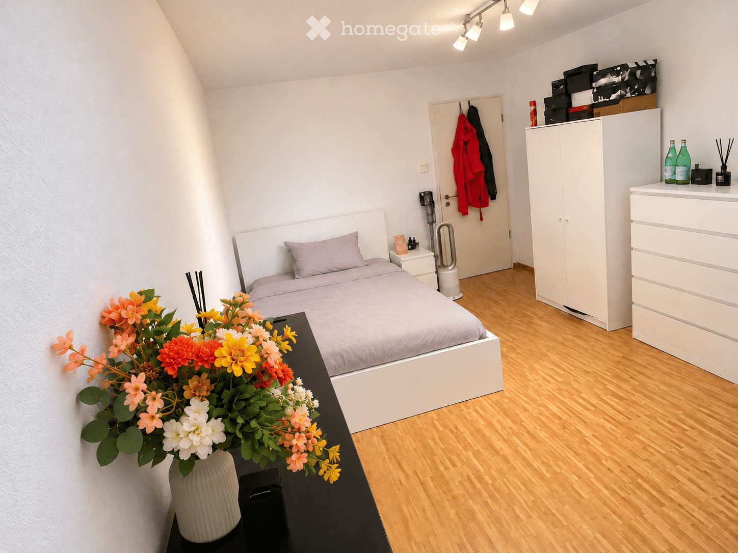 Modern bedroom with a white bed, nightstand, flower vase on a table, wardrobe, and drawer unit. The room features wooden flooring and white walls.