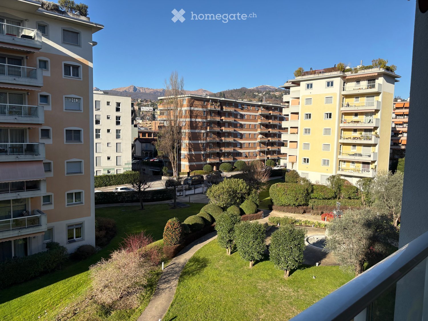 balcony overlooking a green park with shrubs and a walking path, surrounded by multistory apartment buildings