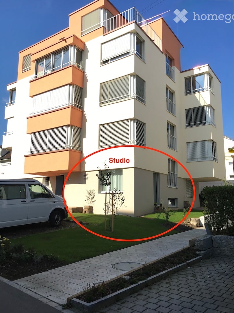 Modern two-story building with white and orange facade, multiple windows, balconies, and a small garden area in front.