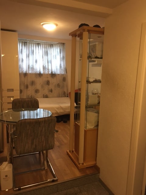 A compact, furnished room with a bed, a small dining area featuring a glass table and two chairs, a mirror cabinet, and a window with curtains.