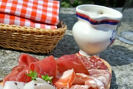 A rustic outdoor setting with a platter of assorted cured meats, a wicker basket with a checkered cloth, and a decorative pitcher.
