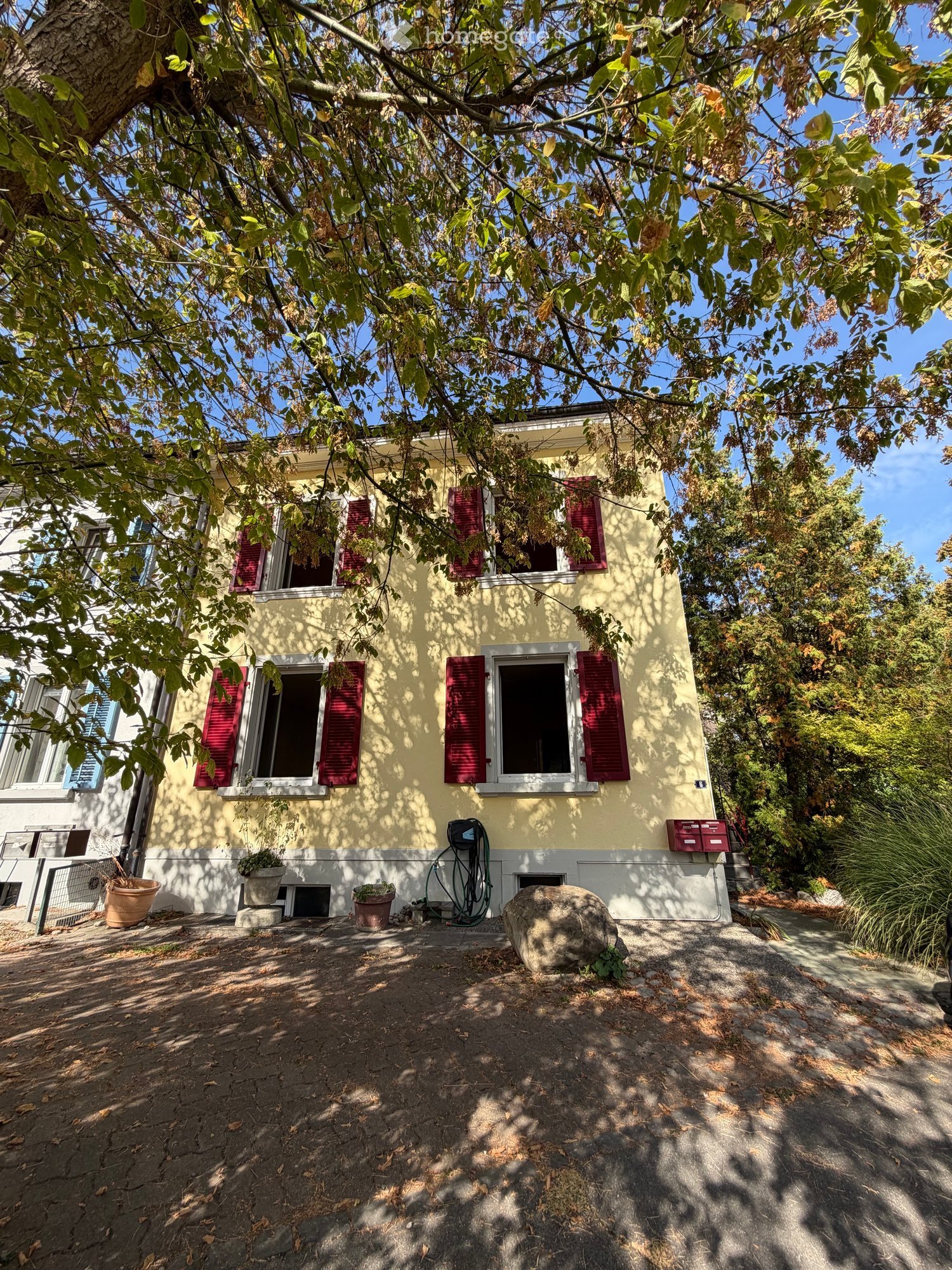 Two-story yellow house with red shutters, surrounded by trees and greenery, with a small garden area and potted plants near the entrance.
