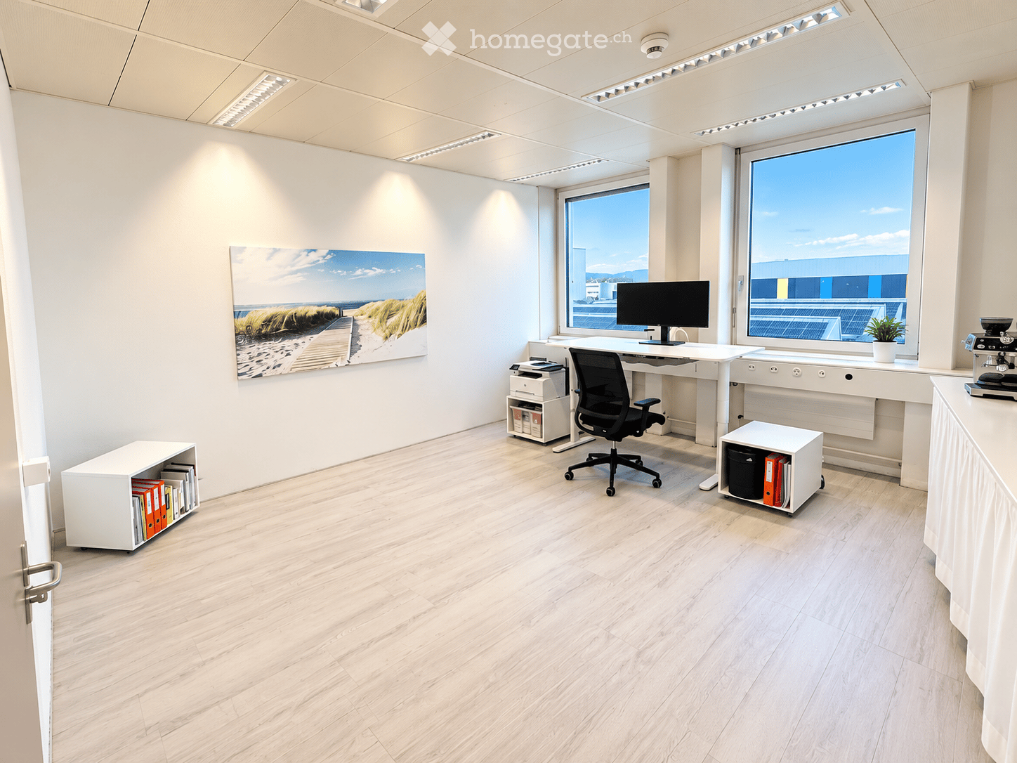 Modern office space with light wood flooring, white walls, large windows, a desk with a chair, a screen, a printer, a cabinet, a coffee machine, a plant, and a picture on the wall.