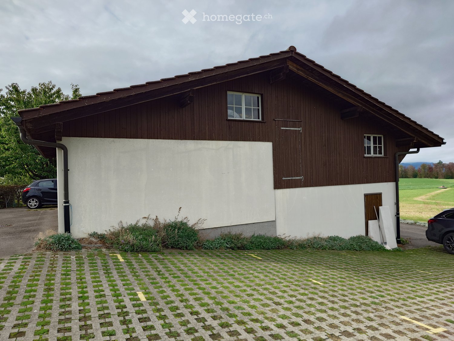 Two-story house with a combination of wooden and white walls, multiple windows, and a tiled roof. The house is surrounded by a paved area with grass patches and has two parked cars nearby.