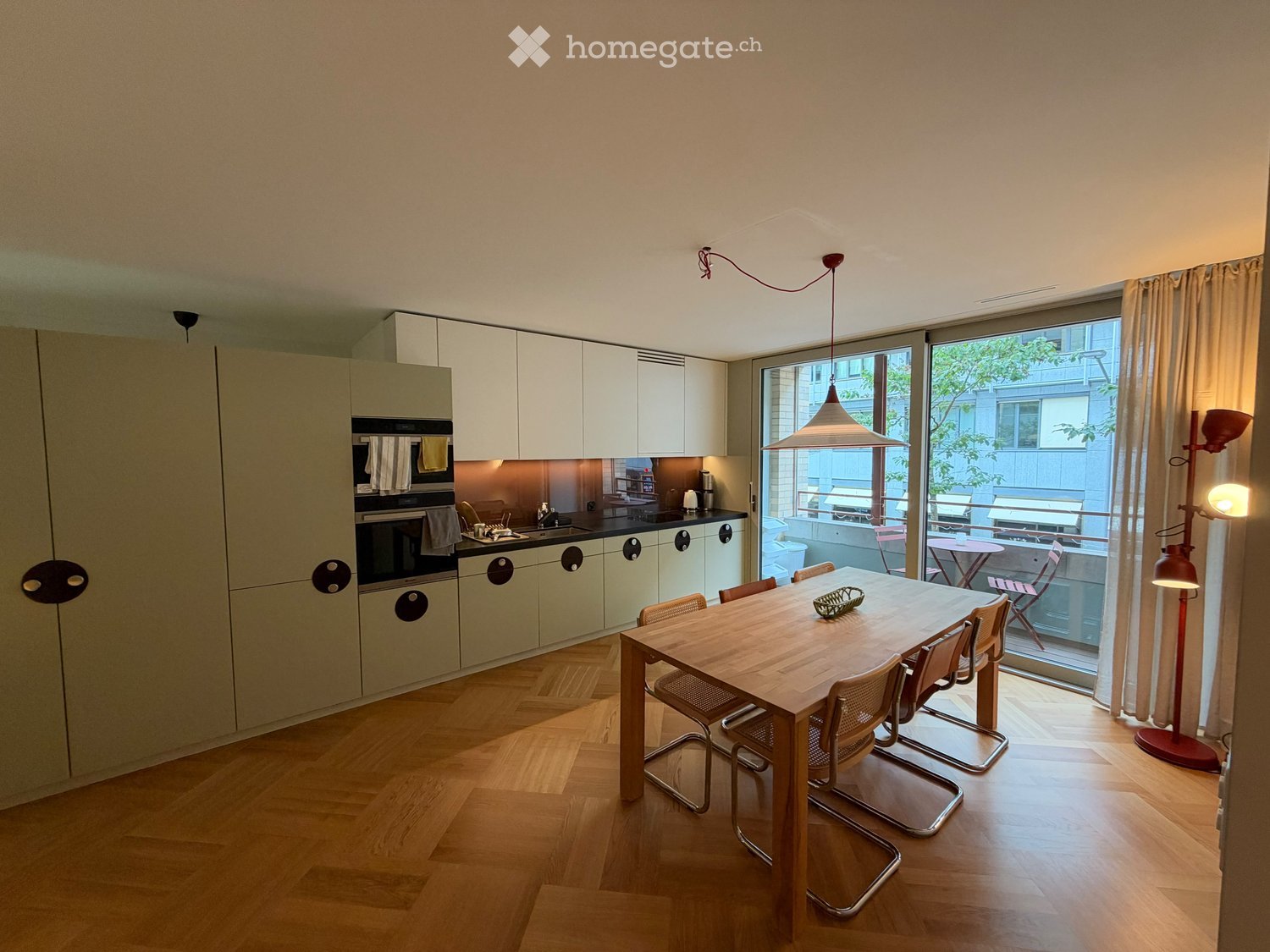 Modern kitchen and dining area with light-colored cabinets, a wooden dining table with six chairs, and a pendant light hanging above the table. The kitchen features an oven, sink, and faucet, with a large window offering a view of the outdoor area.