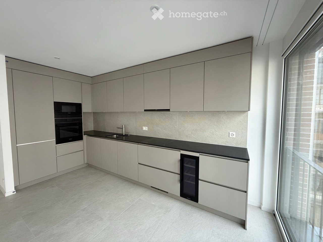 Modern kitchen with light grey cabinets, black countertops, and built-in appliances including an oven and refrigerator. Features a large window with blinds and a sleek, minimalist design.