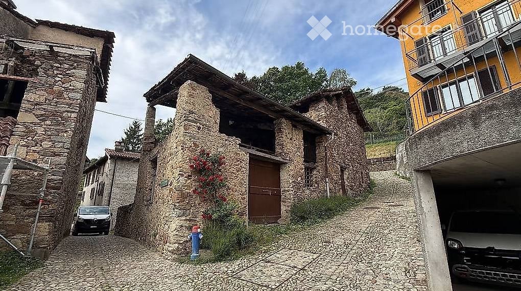 A narrow cobblestone street lined with stone houses, one with a wooden door and small garden, another with a balcony, and a garage with a parked vehicle.