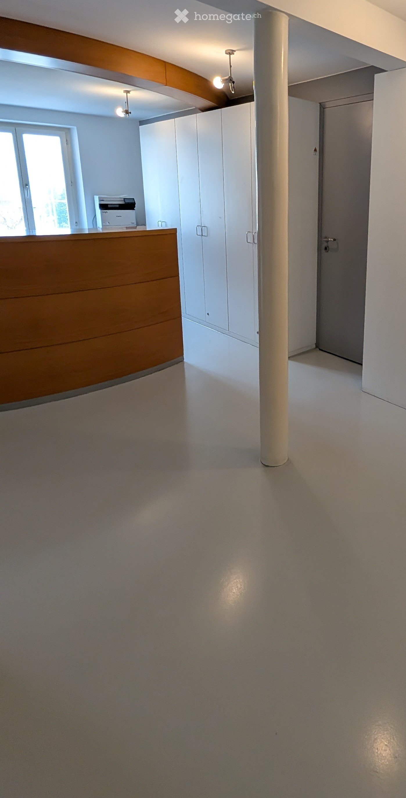 Empty hallway with white walls, a central column, and a wooden reception desk with a printer on top. The space is well-lit with ceiling lights and has a large window on one side.