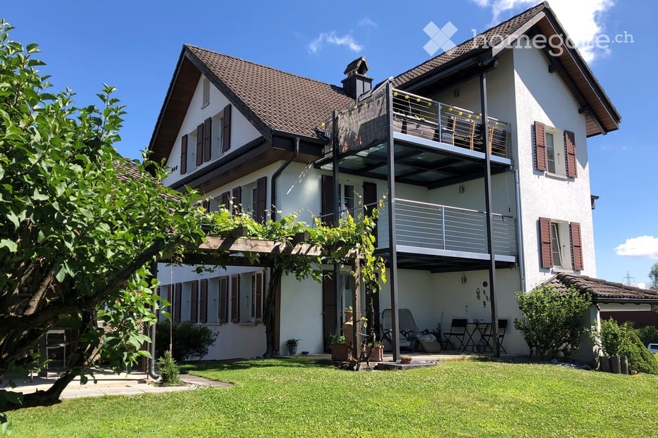 Two-story house with white exterior, brown roof, and balcony on the upper level. The house features multiple windows with shutters, a well-maintained lawn, and outdoor seating area.