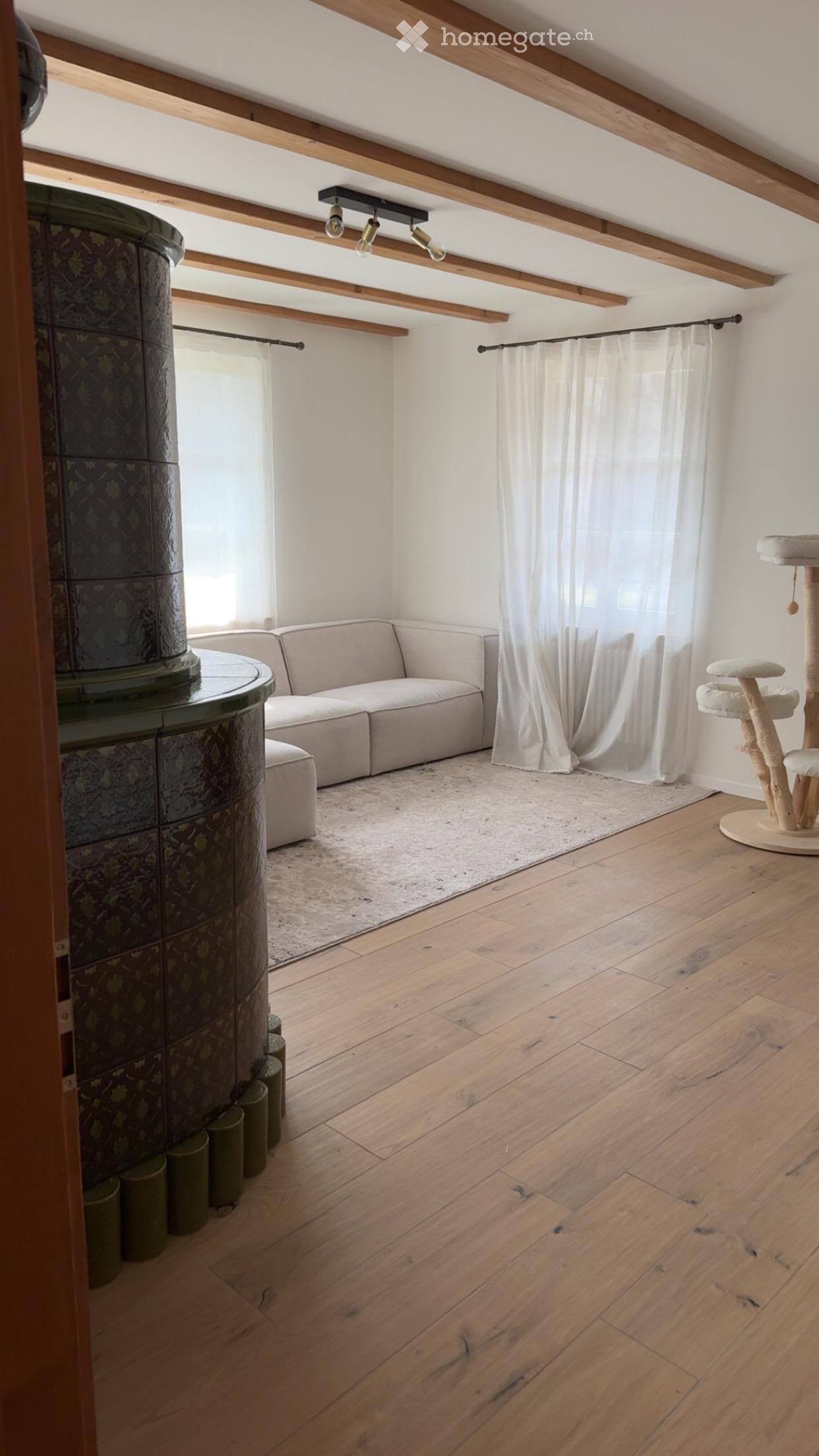 Bright living room with wooden floors, exposed wooden beams, a modern sofa, a cat tree, and a round table with a decorative column.