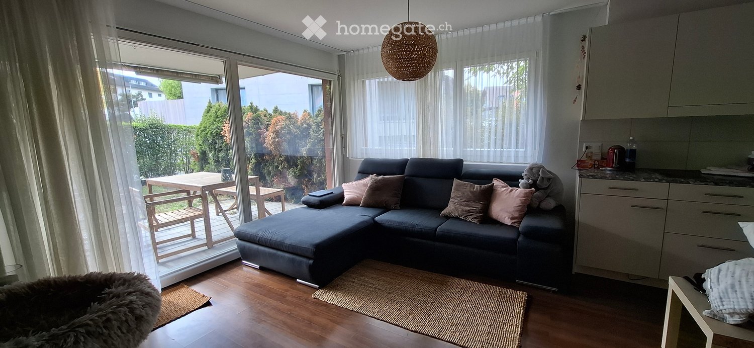 Modern living room with a dark grey sectional sofa, wooden floor, large windows with sheer curtains, and a small kitchen area with cabinets and a counter. A wicker hanging lamp is above the sofa, and a small table and chairs are visible through the glass