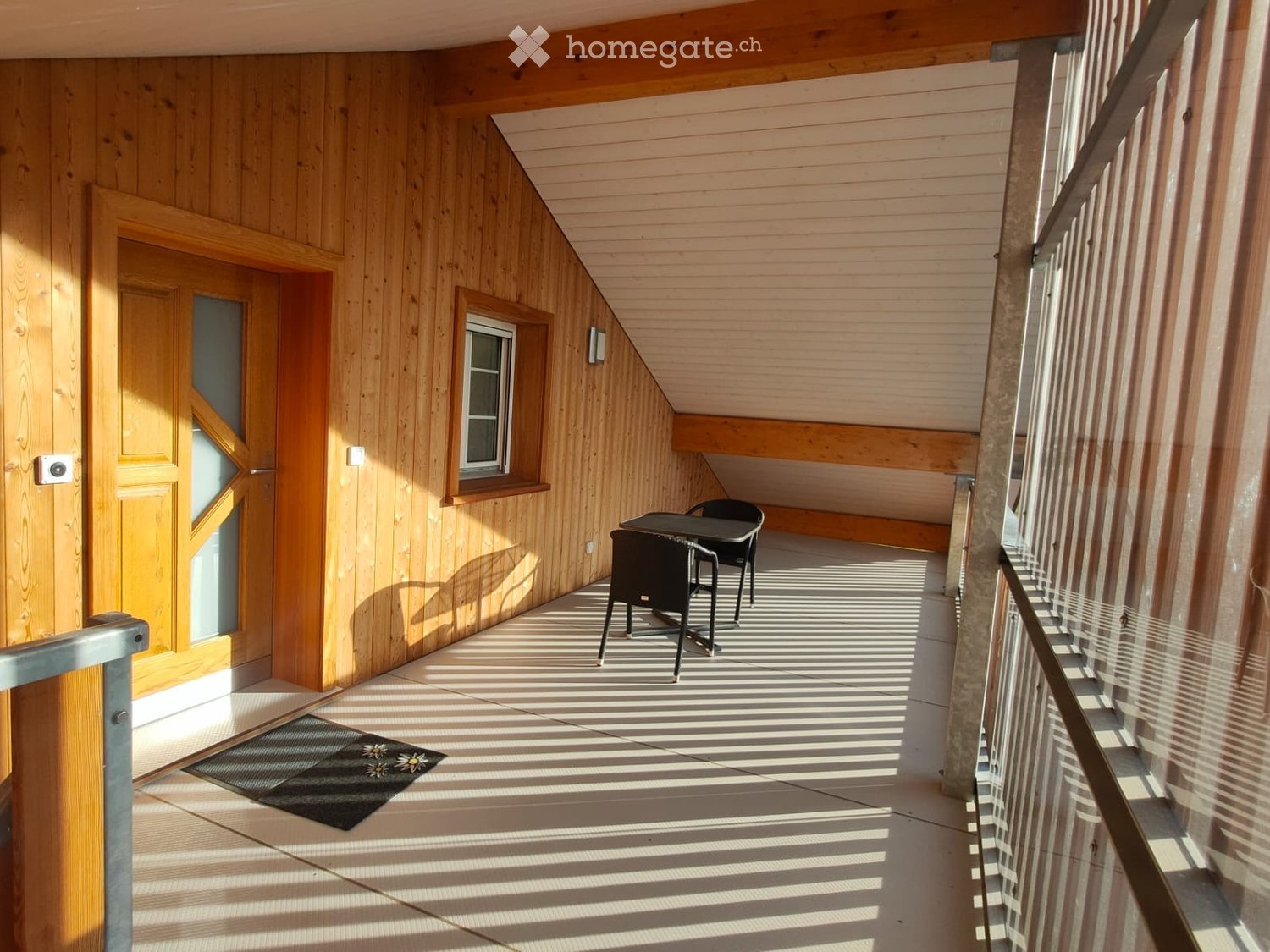 Wooden balcony with a small table and two chairs, wooden walls, a window, and a doormat.