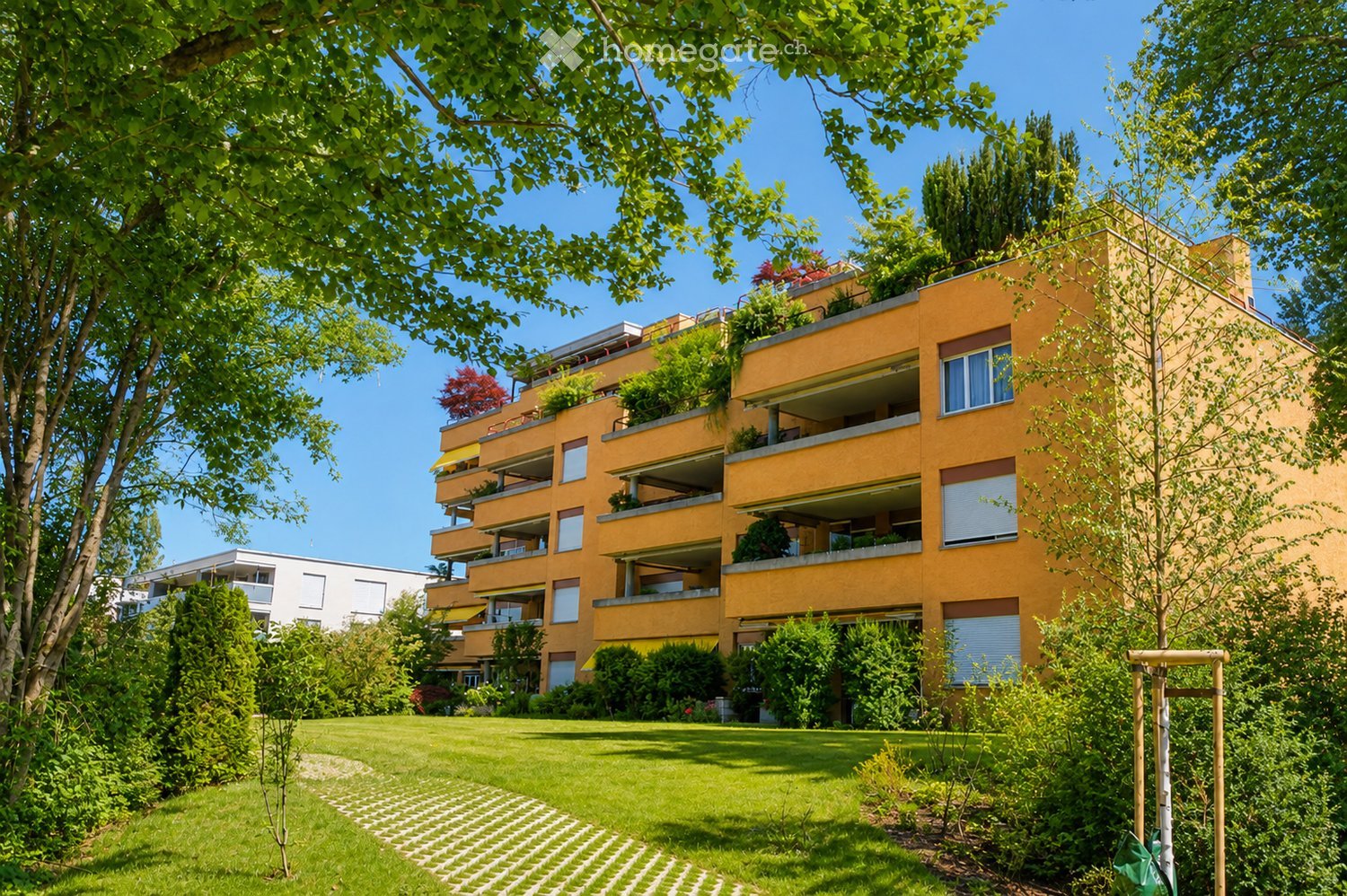 A modern yellow apartment building with multiple balconies and greenery, surrounded by a well-maintained lawn and trees.