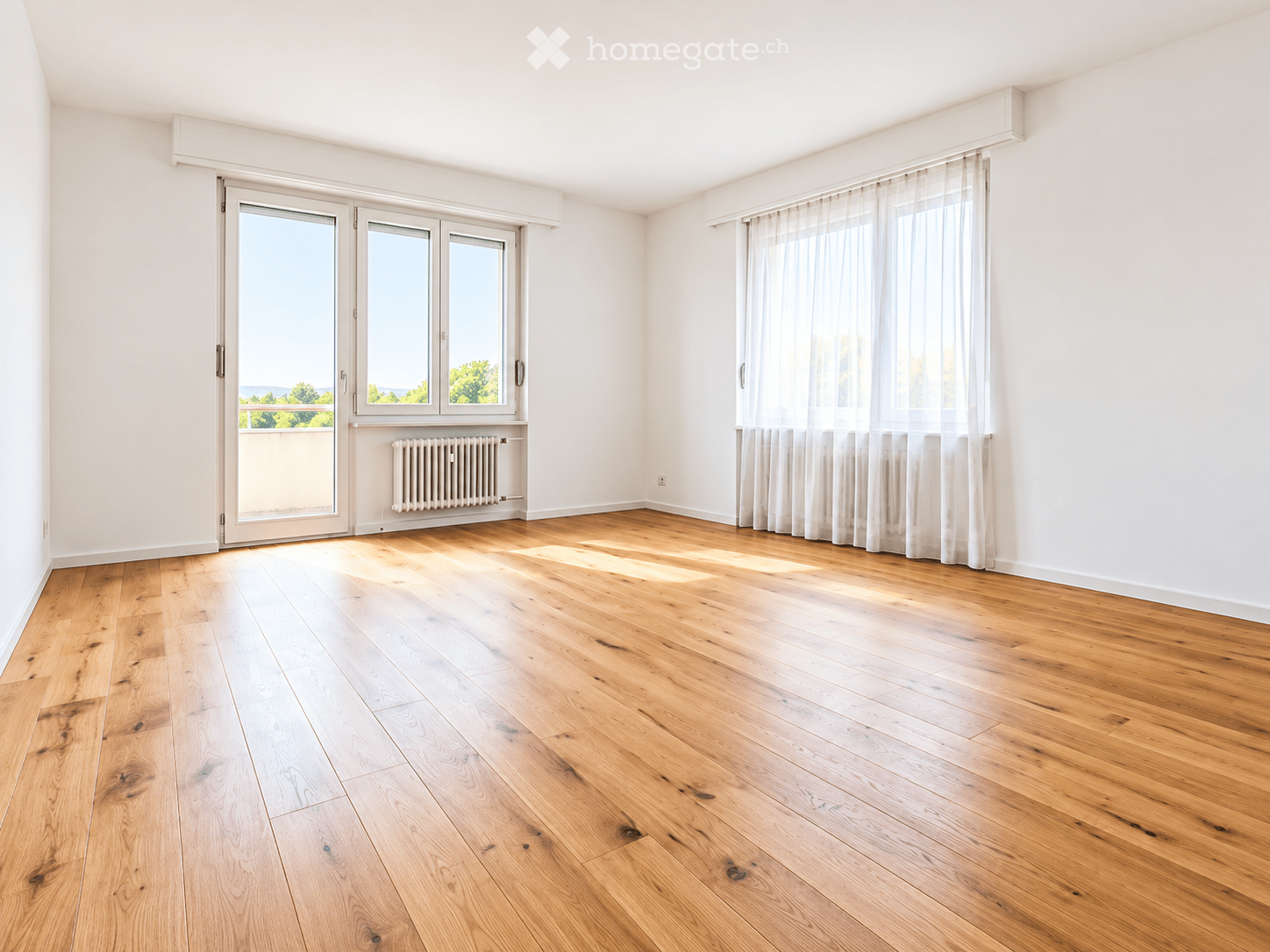 Empty room with light wooden flooring, white walls, large windows with sheer curtains, and a sliding glass door leading to a balcony.