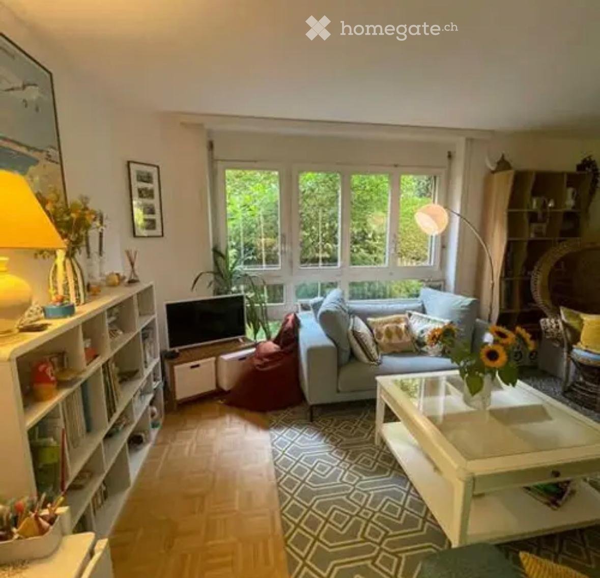 Bright living room with large windows, light grey sofa, coffee table, bookshelf, floor lamp, and decorative items.