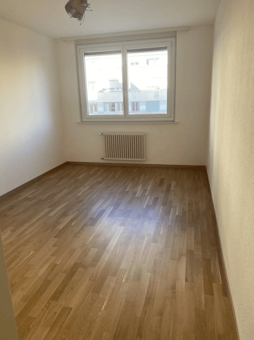 Completely empty room with wooden flooring, white walls, a single window, and a radiator below it.