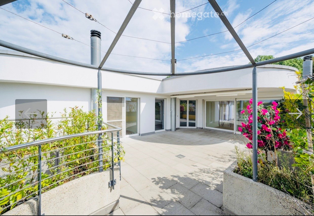 Modern outdoor patio area with white walls, large glass doors, and lush greenery, including flowering plants and shrubs.