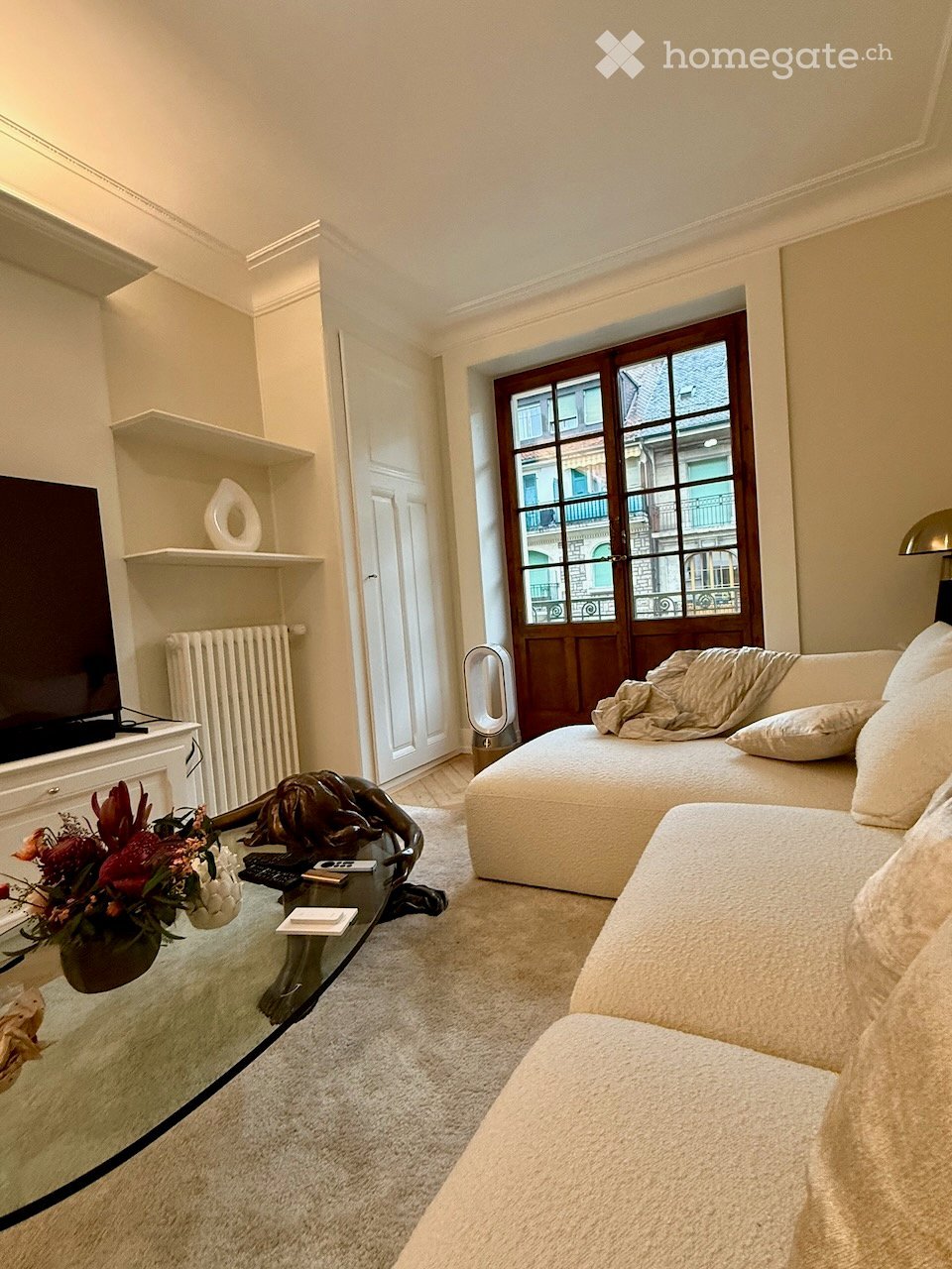 Modern living room with a white sofa, glass coffee table, decorative sculpture, flower arrangement, TV on a white cabinet, and large windows with wooden frames.