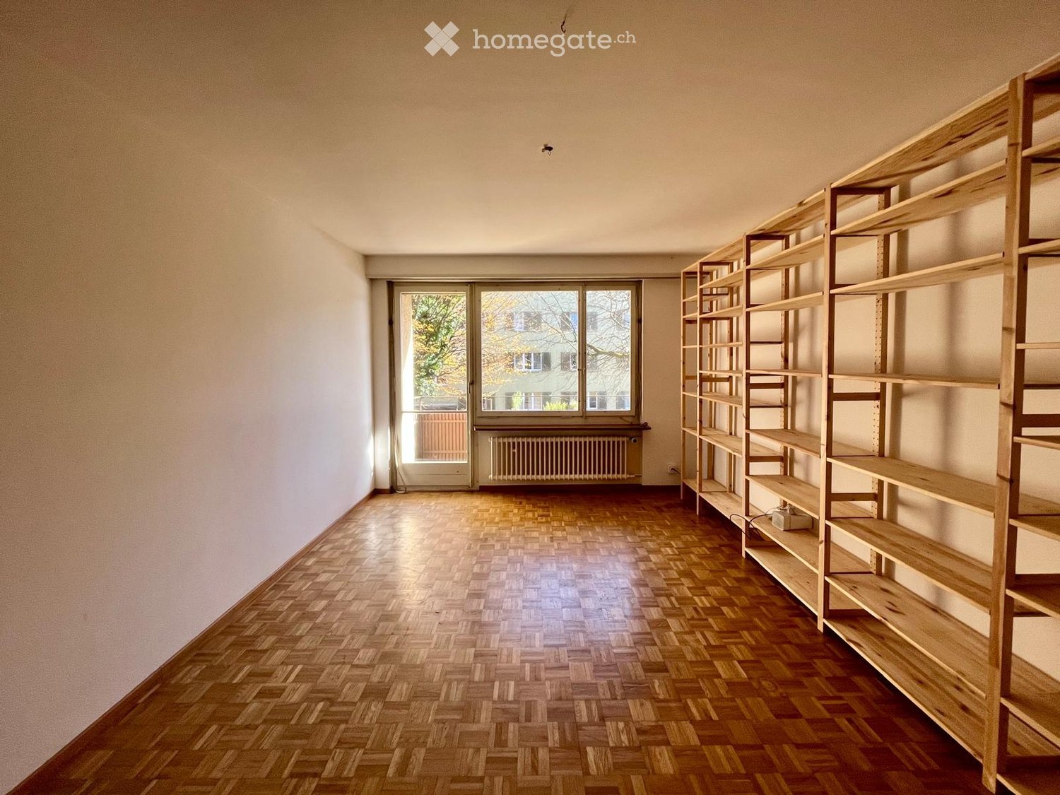 Empty room with wooden herringbone flooring, white walls, large windows, and built-in wooden shelving units.