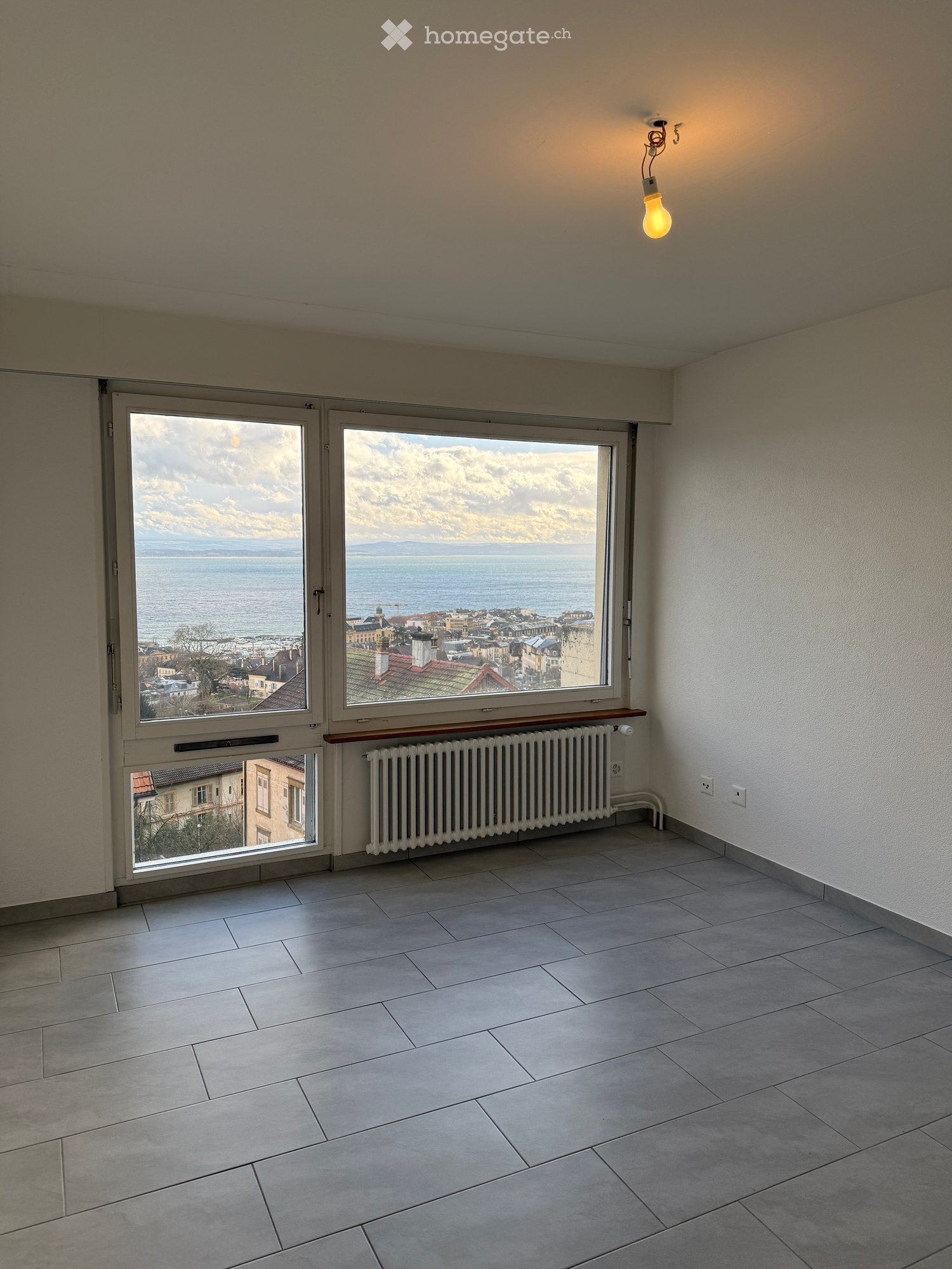 Empty room with tiled floor, large windows offering a scenic view of a city and body of water, radiator below the windows, and a single light fixture on the ceiling.