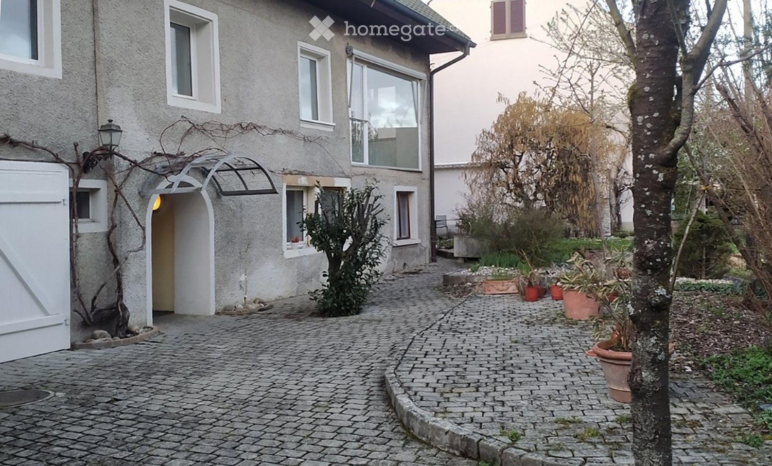 A cobblestone pathway leads to a house entrance with a small garden area featuring potted plants and trees.