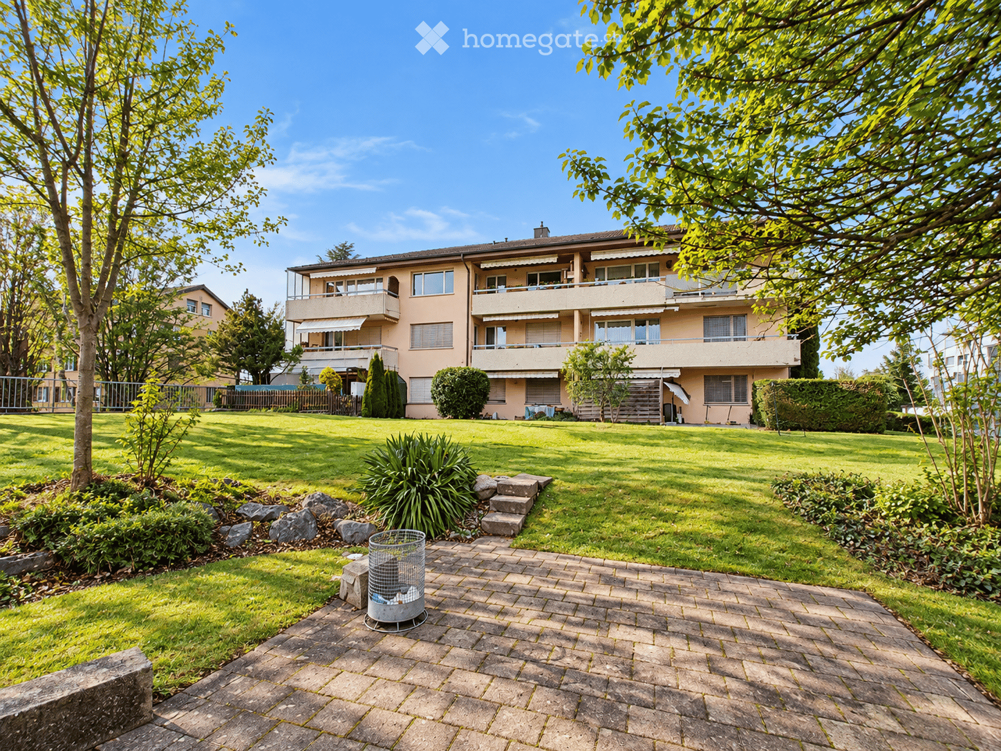 Well-maintained garden with a paved pathway, grassy lawn, shrubs, trees, and a multi-story residential building with balconies in the background.