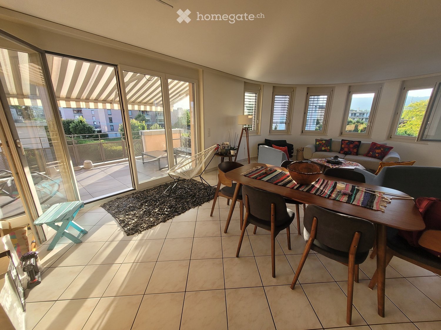 Bright living room with large windows and a balcony, featuring a sofa, dining table, chairs, and a rug.
