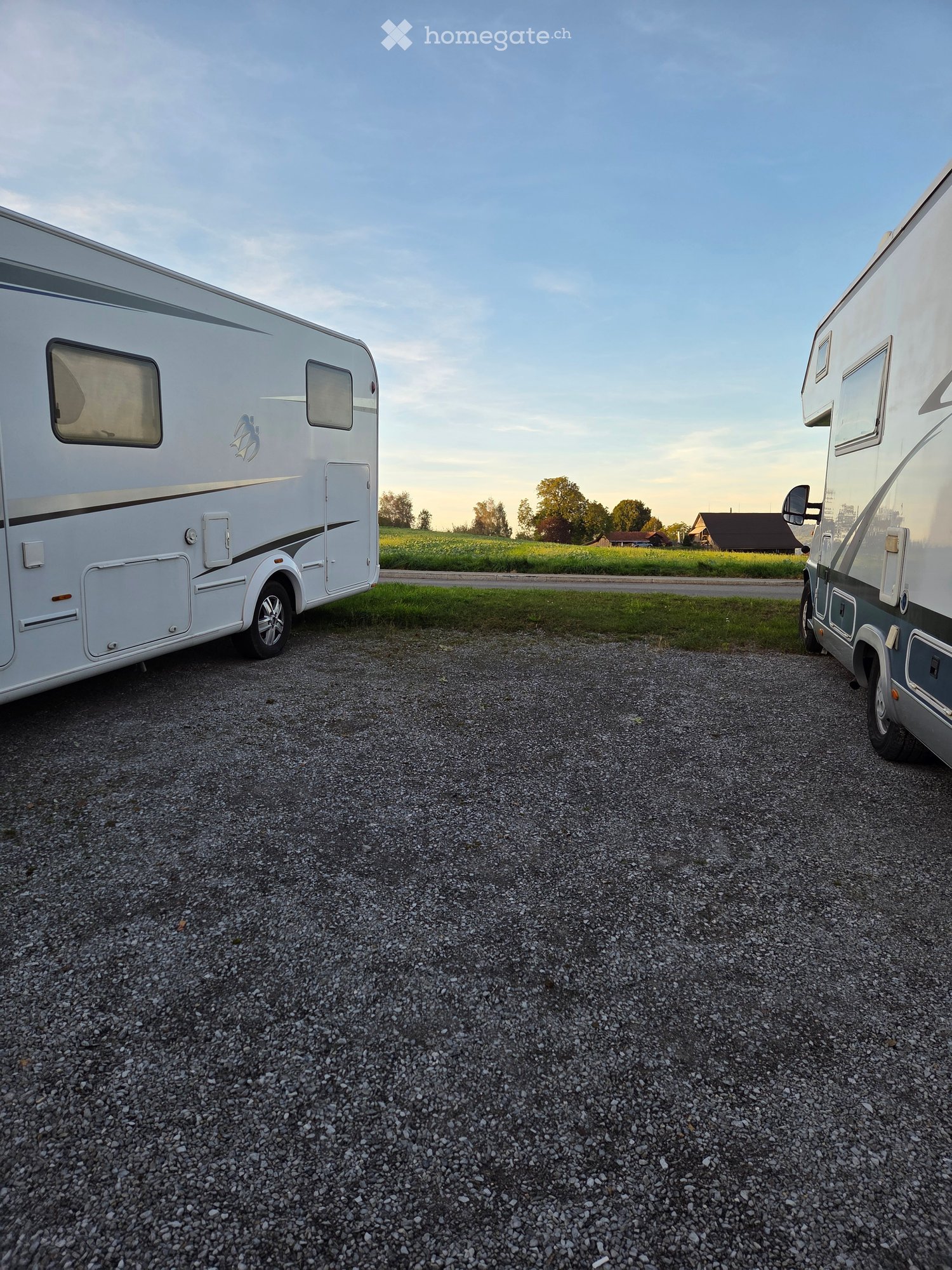 Two white RVs parked on a gravel surface in a rural area with green fields and scattered houses in the background.