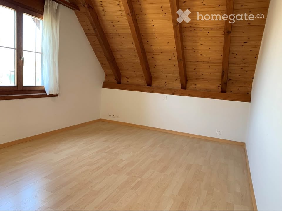 Empty room with wooden flooring, white walls, and exposed wooden beams on the ceiling. A window with white curtains is visible on one side.