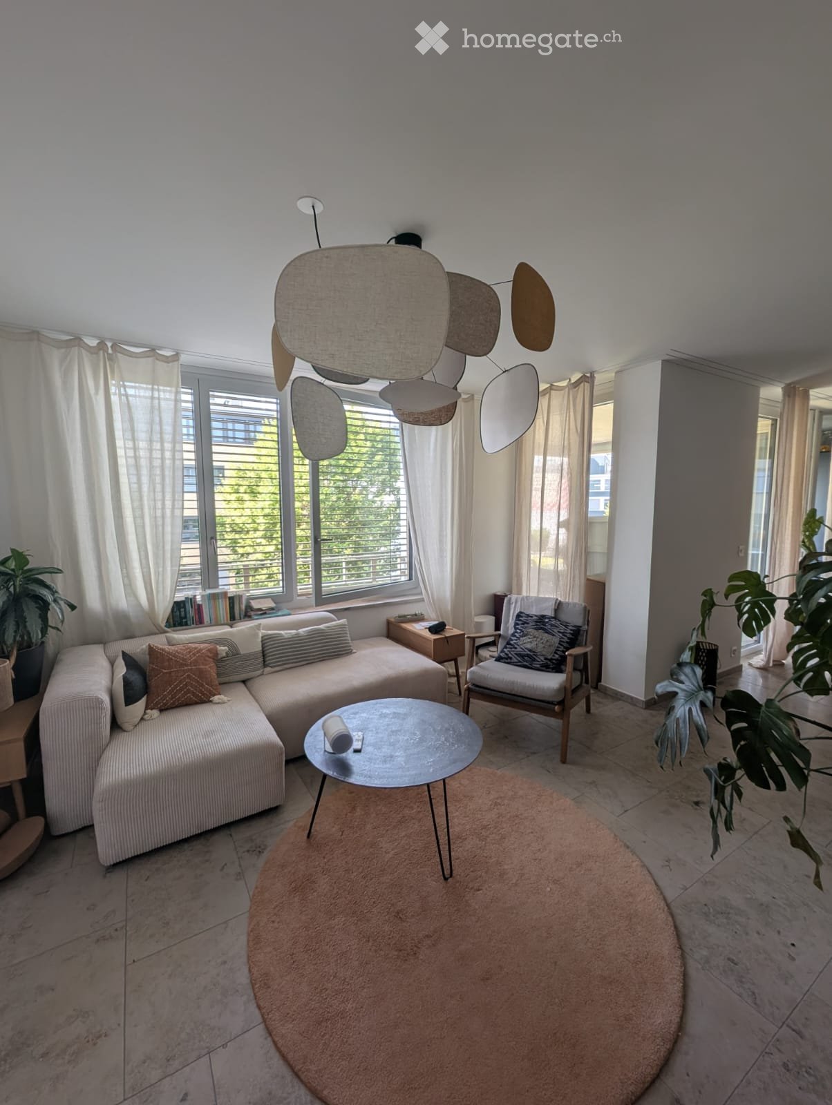 Modern living room with a grey sectional sofa, wooden coffee table, armchair, and decorative floor lamp. Large windows with white curtains and plants add to the bright ambiance.