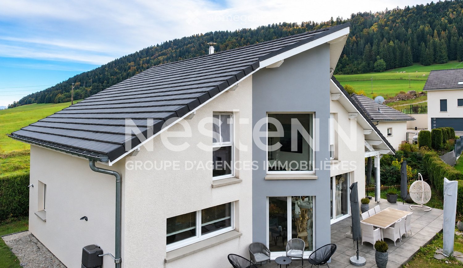 Modern two-story house with grey and white exterior, large windows, and a spacious outdoor patio with furniture and a scenic mountain view.