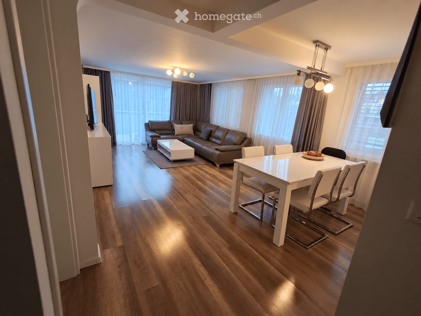 Modern living and dining area with wooden flooring, large windows with curtains, a brown leather sofa, a white coffee table, a white dining table with six chairs, and a TV on a white stand.