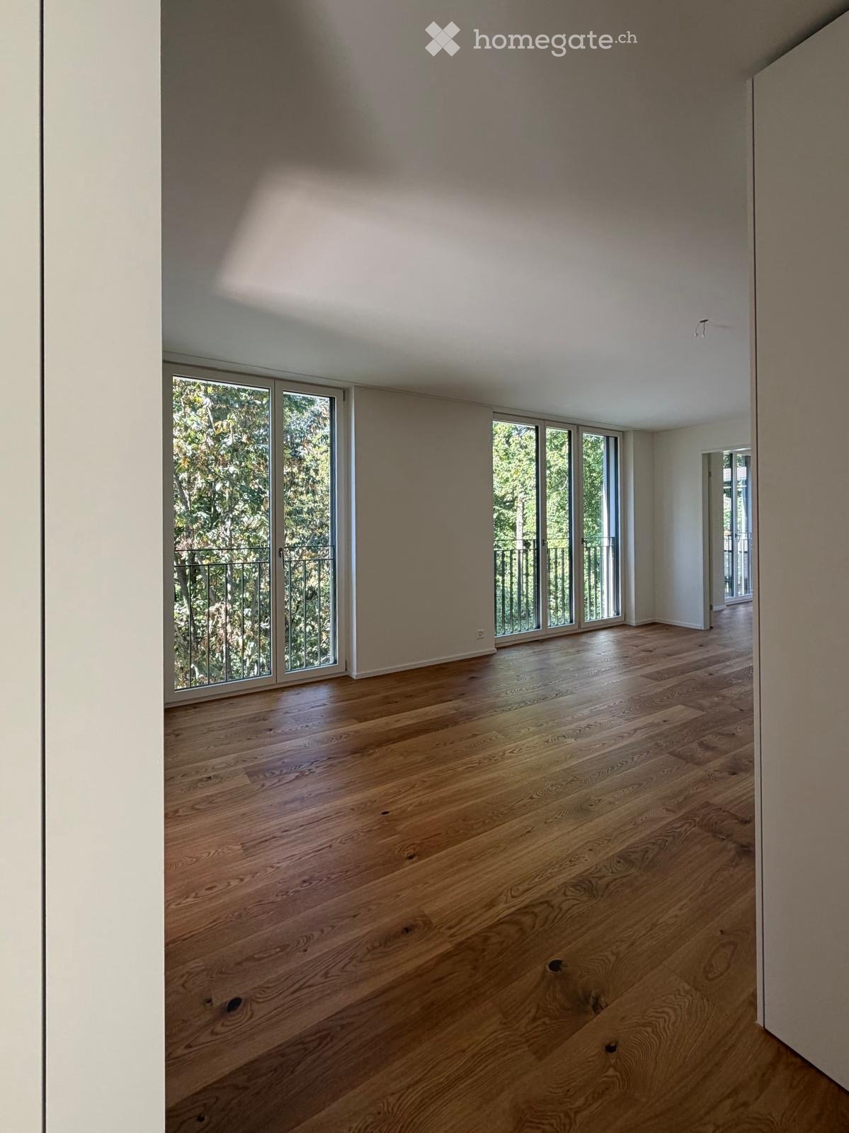Empty room with wooden flooring, large windows, and a view of greenery outside. No furniture or equipment present.