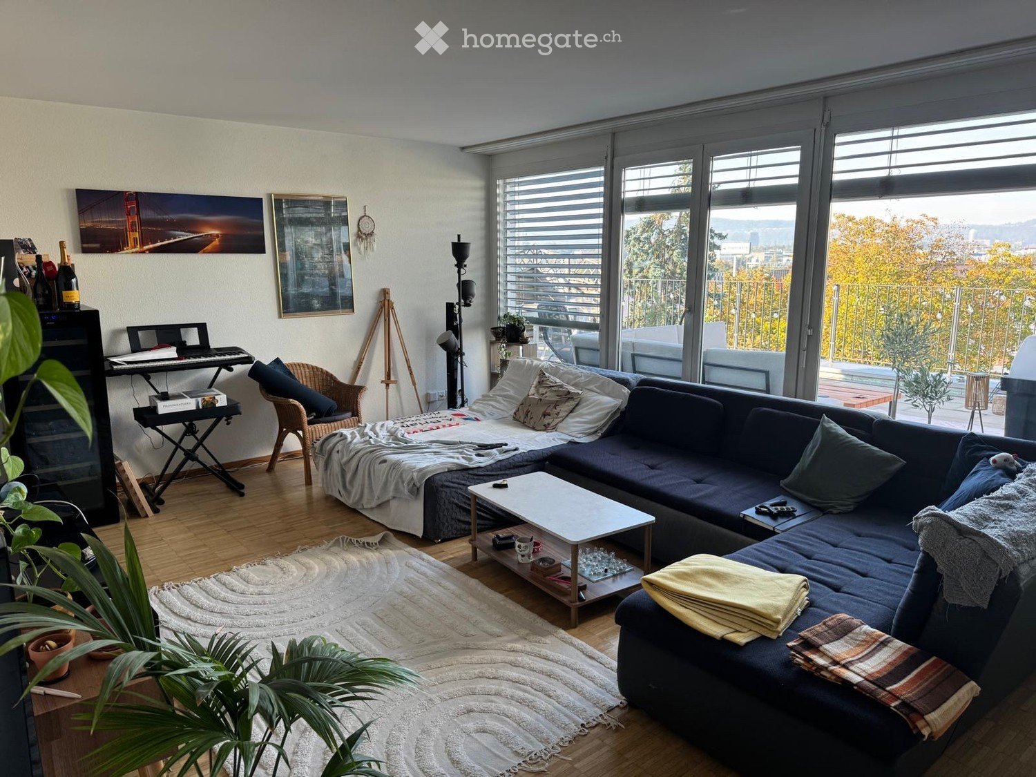 Modern living room with large windows offering a scenic view, featuring a dark blue sectional sofa, a white coffee table, a wicker chair, a small keyboard, and decorative plants.