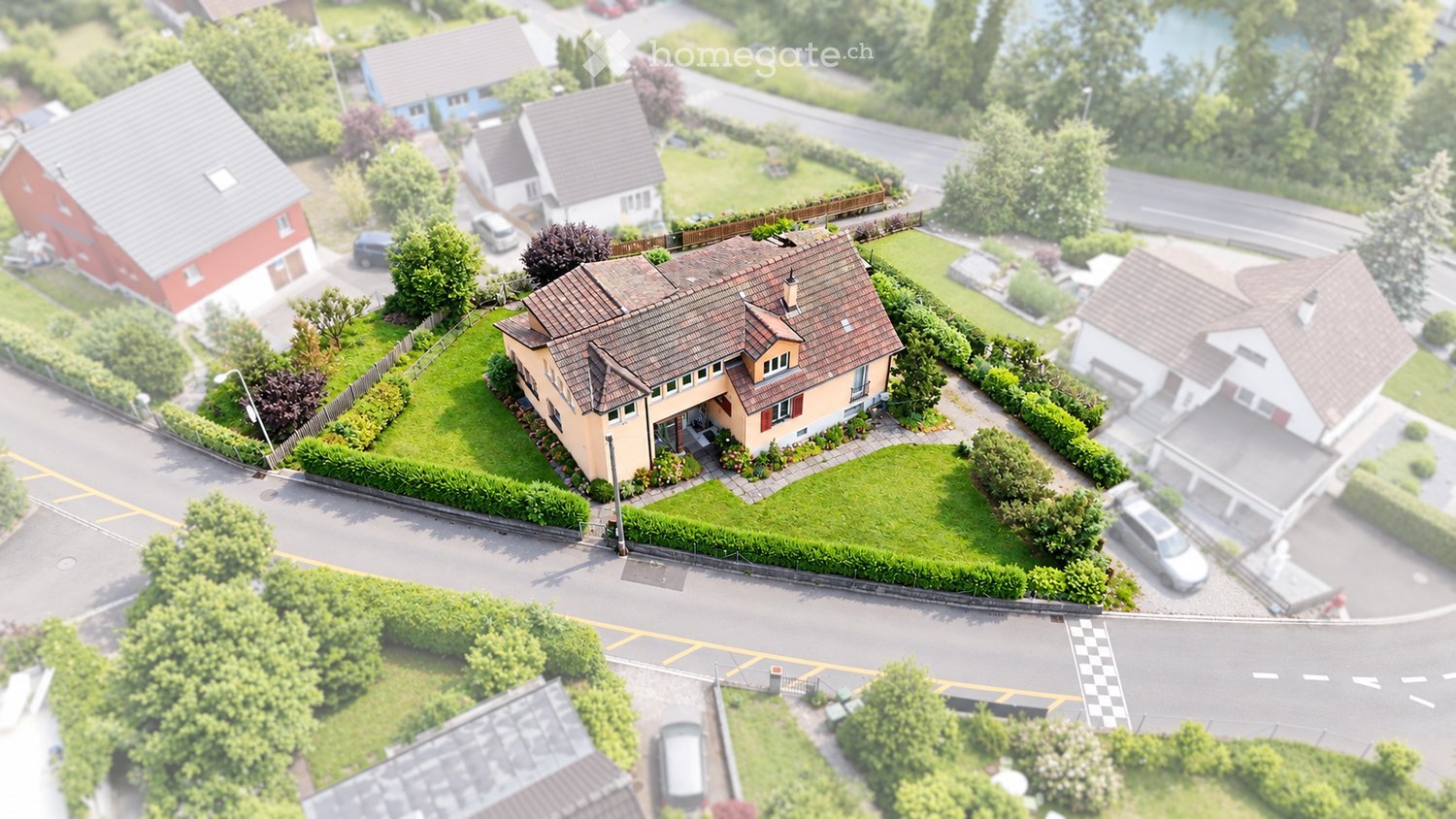 Aerial view of a two-story house with a brown tiled roof, surrounded by a well-maintained garden and a driveway with parked cars.