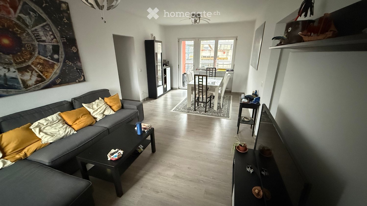 Open-plan living and dining area with a grey sectional sofa, coffee table, dining table with chairs, and a large window leading to a balcony.